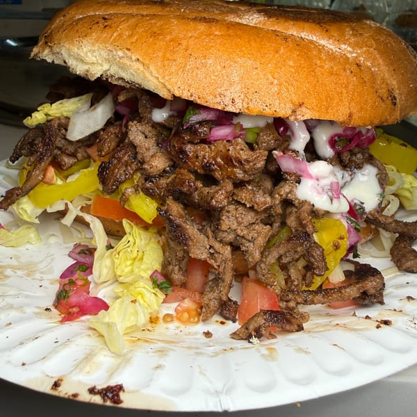 Loaded grilled meat sandwich on toasted bread with lettuce, tomatoes, peppers, pickled onions, and creamy sauce.